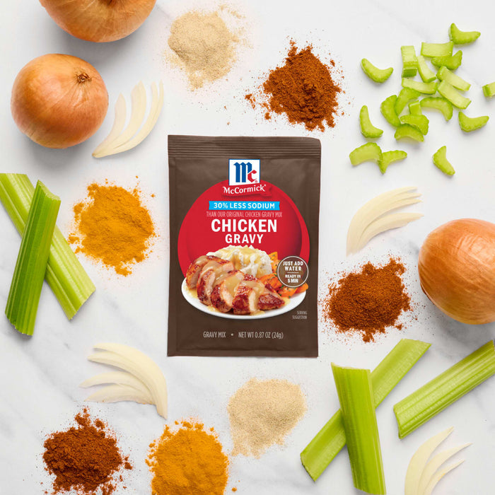A packet of McCormick Chicken Gravy mix is surrounded by fresh ingredients like onions, celery, garlic, and spices, suggesting a homemade gravy recipe.