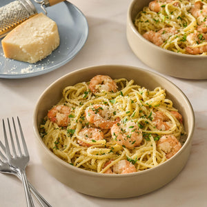 Two bowls of pasta are topped with shrimp and fresh herbs prepared using Mccormick Garlic Butter Shrimp Scampi Seasoning Mix .87 Oz alongside a block of parmesan cheese.