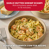 A hand pours a packet of Mccormick Garlic Butter Shrimp Scampi Seasoning Mix .87 Oz over a skillet filled with cooked pasta and shrimp next to a wedge of parmesan cheese.