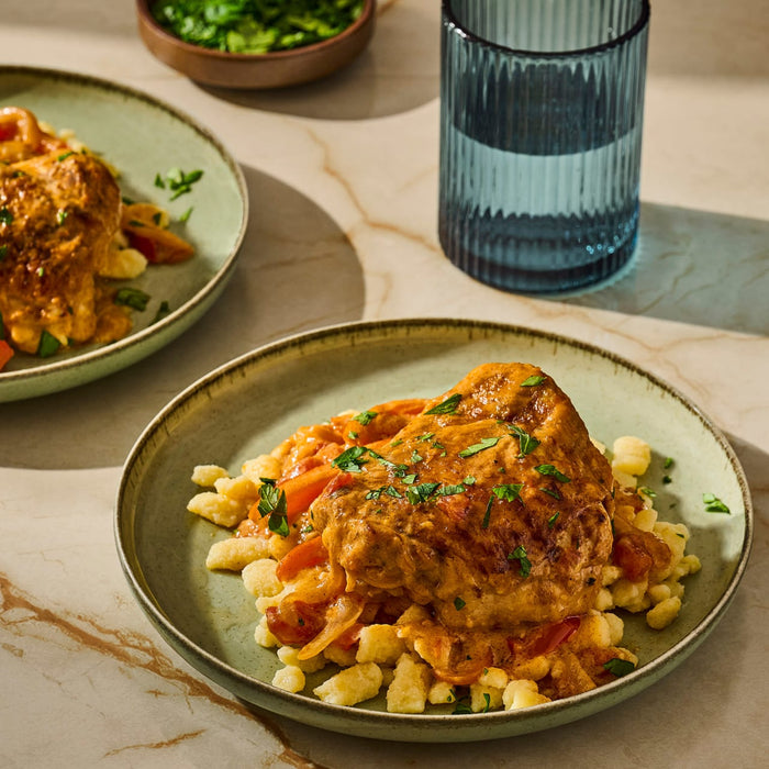 A delicious plate of chicken paprikash with spaetzle is presented alongside a glass of water and a bowl of greens.