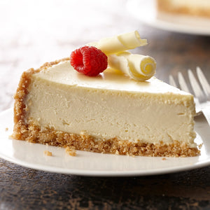 A slice of cheesecake topped with a raspberry and white chocolate curls sits on a white plate.