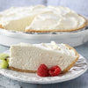A slice of creamy cheesecake is served on a plate with raspberries and kiwi with the rest of the pie in the background.