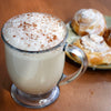 A glass mug filled with a frothy beverage sprinkled with cinnamon sits next to a plate of frosted cinnamon rolls.