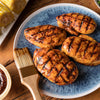 Grilled chicken breasts with barbecue sauce are arranged on a blue plate, alongside corn on the cob and a brush.