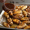 A batch of cooked chicken wings on a baking sheet is being drizzled with a flavorful sauce.
