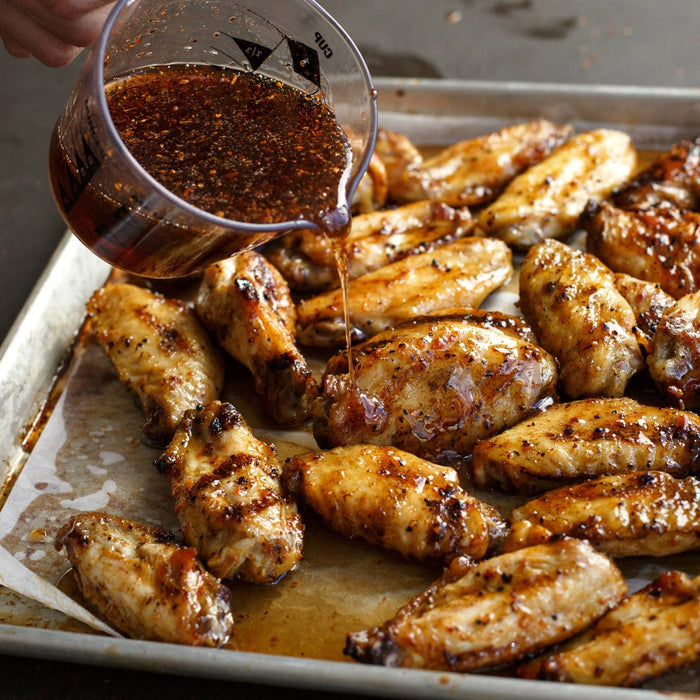 A batch of cooked chicken wings on a baking sheet is being drizzled with a flavorful sauce.