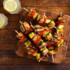 A rustic display of grilled chicken and vegetable skewers, prepared with Mccormick Grill Mates Mesquite Seasoning 24 Oz, rests on a wooden board alongside two glasses of lemon water.