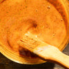 A close-up shows a bowl of orange sauce with herbs being stirred with a wooden brush.