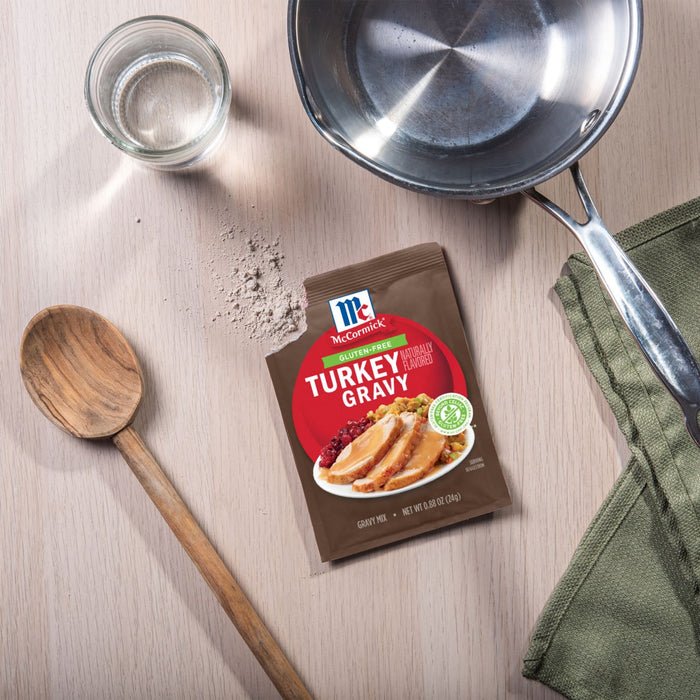 A packet of McCormick Turkey Gravy mix sits on a wooden surface next to a pot, a glass of water, a wooden spoon, and a green cloth.
