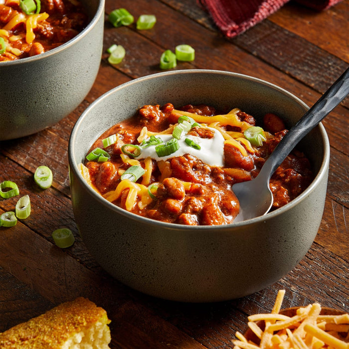 Two bowls of chili topped with shredded cheese, sour cream, and green onions are displayed on a wooden surface, suggesting a hearty and comforting meal.