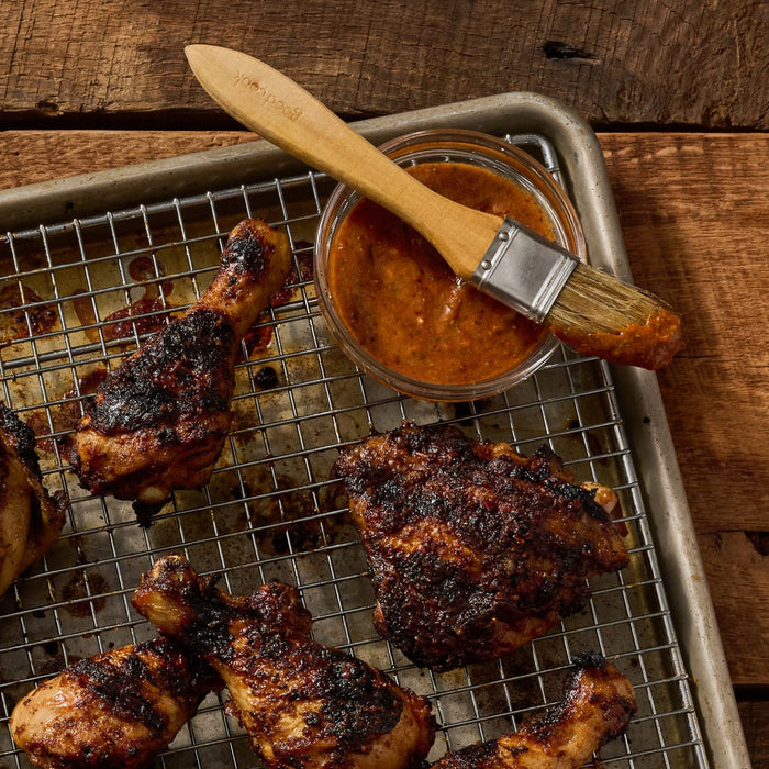 A baking sheet with a wire rack holds grilled chicken drumsticks and thighs, alongside a bowl of sauce and a basting brush, all prepared with Mccormick Grill Mates Brown Sugar Bourbon Blend Gf 3.0 Oz.