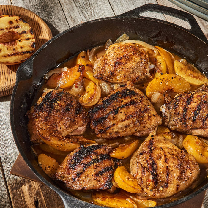 A cast iron skillet holds grilled chicken thighs, caramelized peaches, and onions, all seasoned with Mccormick Grill Mates Brown Sugar Bourbon Blend Gf 3.0 Oz, with a grilled peach half on a wooden board beside it.