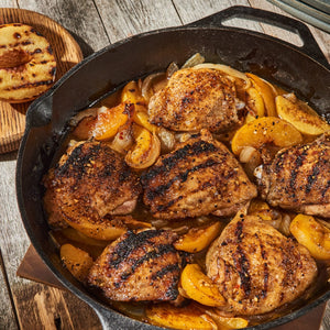A cast iron skillet holds grilled chicken thighs with peaches and onions, sitting on a wooden table with a grilled peach half on a wooden board.