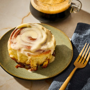 A frosted cinnamon roll on a green plate next to a cup of coffee and a gold fork.