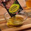 A hand pours McCormick Grill Mates Brazilian Steakhouse marinade mix into a bowl of oil.