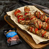 Grilled chicken and red pepper skewers are displayed on a wooden board next to a bag of Jack Link's Montreal Steak jerky.