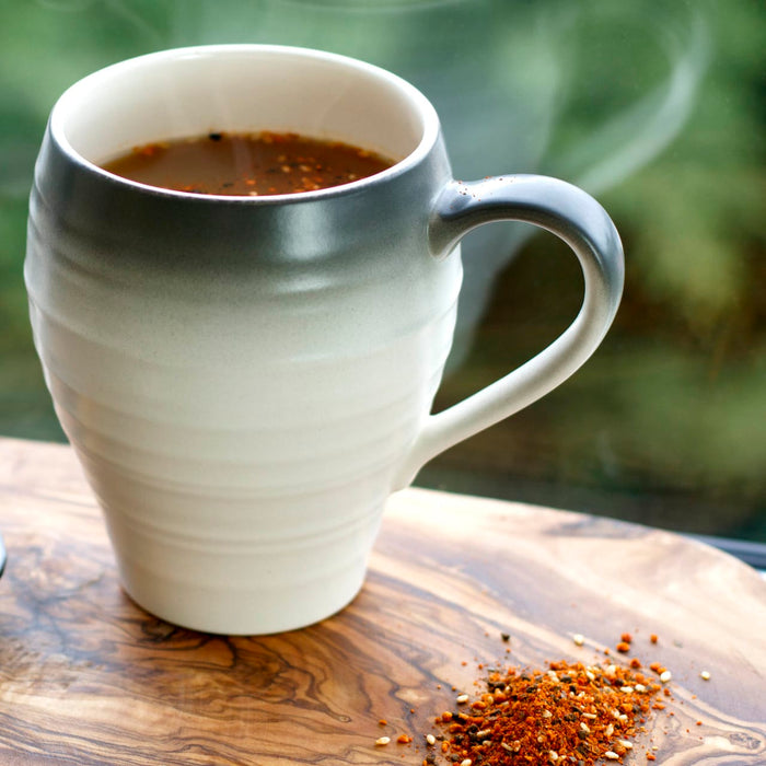 A white mug with a grey rim filled with a dark liquid sits on a wooden cutting board with a pile of red and white spices.