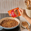 A hand pours Mccormick Taco Seasoning Mix from its orange packet onto cooked ground meat in a black pan.