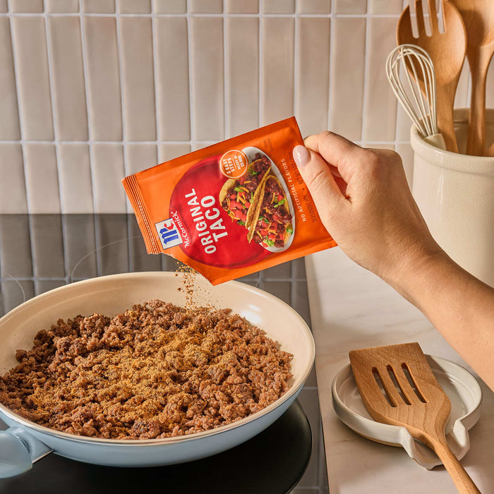 A hand pours Mccormick Taco Seasoning Mix from its orange packet onto cooked ground meat in a black pan.