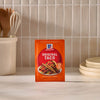 A package of Mccormick Taco Seasoning Mix is displayed on a wooden table alongside two plates and a baking dish filled with a hearty taco casserole, featuring ground meat, corn, beans, and a topping of golden tater tots.
