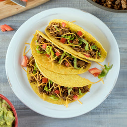 Three tacos filled with ground beef shredded cheese lettuce and diced tomatoes on a white plate.