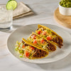 A plate of three hard-shell tacos, prepared with Mccormick Taco Seasoning Mix, are filled with seasoned ground beef, shredded lettuce, diced tomatoes, and shredded cheese, with guacamole and cooking ingredients visible in the background.