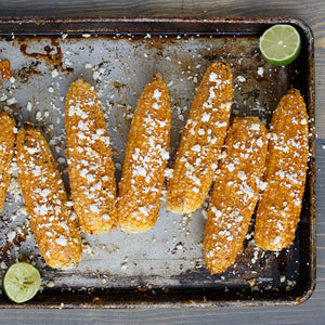A baking sheet with grilled corn on the cob topped with cotija cheese and lime wedges.