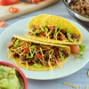 Two tacos filled with ground beef shredded cheese lettuce and diced tomatoes on a white plate.