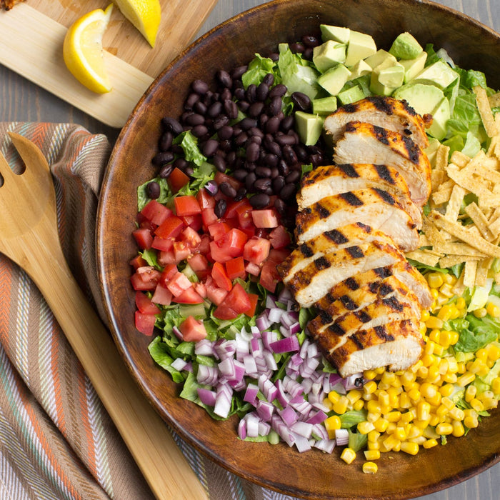 A wooden bowl filled with a salad with grilled chicken black beans corn avocado red onion tomatoes and tortilla strips.