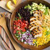 A wooden bowl filled with a salad with grilled chicken black beans corn avocado red onion tomatoes and tortilla strips.