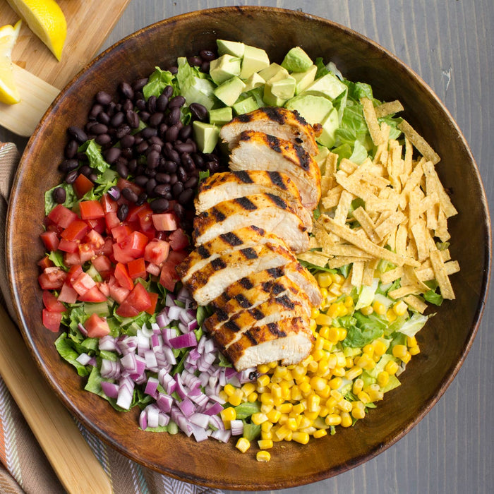 A wooden bowl filled with a salad with grilled chicken black beans corn avocado tomatoes red onion lettuce and tortilla strips.