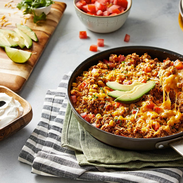 A skillet of cheesy quinoa with ground beef corn tomatoes and avocado slices.