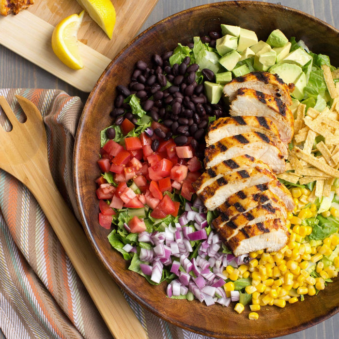 A wooden bowl filled with a salad with grilled chicken black beans corn avocado tomatoes red onion and tortilla strips.