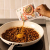 A person pours a packet of Mccormick Organics Regular Taco Seasoning Mix 1 Oz onto cooked ground meat in a white skillet to prepare taco filling.