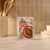 A packet of Mccormick Organics Regular Taco Seasoning Mix 1 Oz is set on a kitchen countertop, with a crock of wooden utensils and a stack of plates in the background.