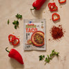 A flat lay image shows a packet of Mccormick Organics Regular Taco Seasoning Mix 1 Oz surrounded by fresh ingredients like red peppers, oregano, and a pile of the seasoning on a marble countertop.