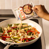 A hand pours a packet of Mccormick Organics Regular Fajita Seasoning Mix 1 Oz over sizzling chicken, onions, and peppers in a white skillet.