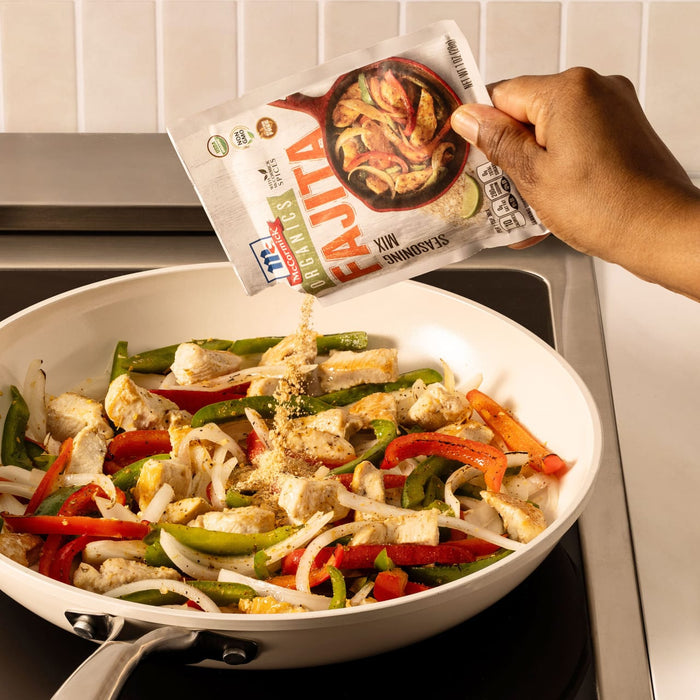 A hand pours a packet of Mccormick Organics Regular Fajita Seasoning Mix 1 Oz over sizzling chicken, onions, and peppers in a white skillet.