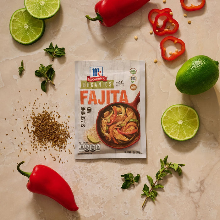 A packet of Mccormick Organics Regular Fajita Seasoning Mix 1 Oz is displayed on a countertop, artfully arranged with fresh ingredients like red peppers, limes, oregano, and spices to suggest a delicious meal.
