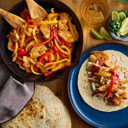A top-down view shows a delicious meal of chicken fajitas, made using Mccormick Organics Regular Fajita Seasoning Mix 1 Oz, served from a cast-iron skillet onto tortillas with fresh limes and a drink.