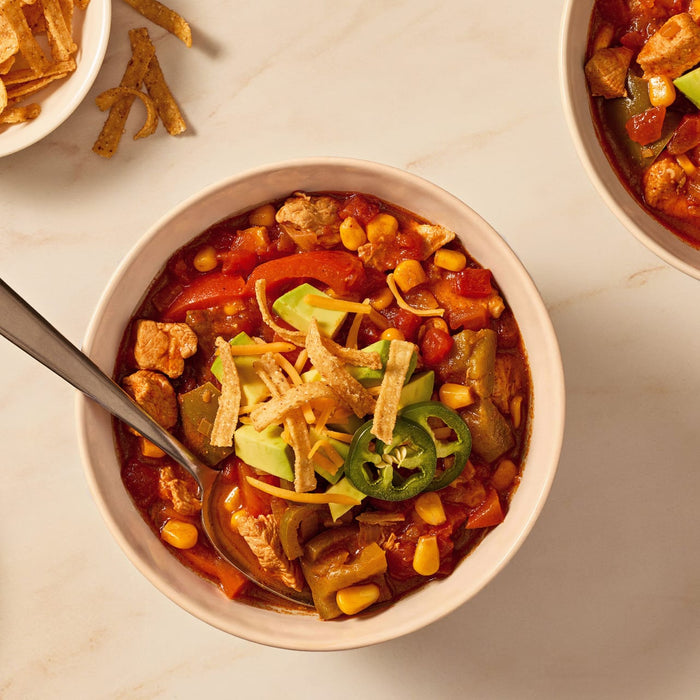 A hearty bowl of chicken fajita soup, made with Mccormick Organics Regular Fajita Seasoning Mix 1 Oz, is garnished with fresh avocado, tortilla strips, and a slice of jalapeño.