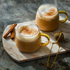 Two mugs of chai latte with frothy milk and cinnamon on a marble board with cinnamon sticks and a tea infuser.