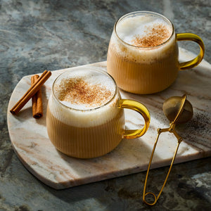 Two mugs of chai latte with frothy milk and cinnamon on a marble board with cinnamon sticks and a tea infuser.