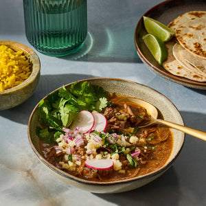 A bowl of hearty stew garnished with radishes and cilantro, accompanied by yellow rice, tortillas, and lime wedges.