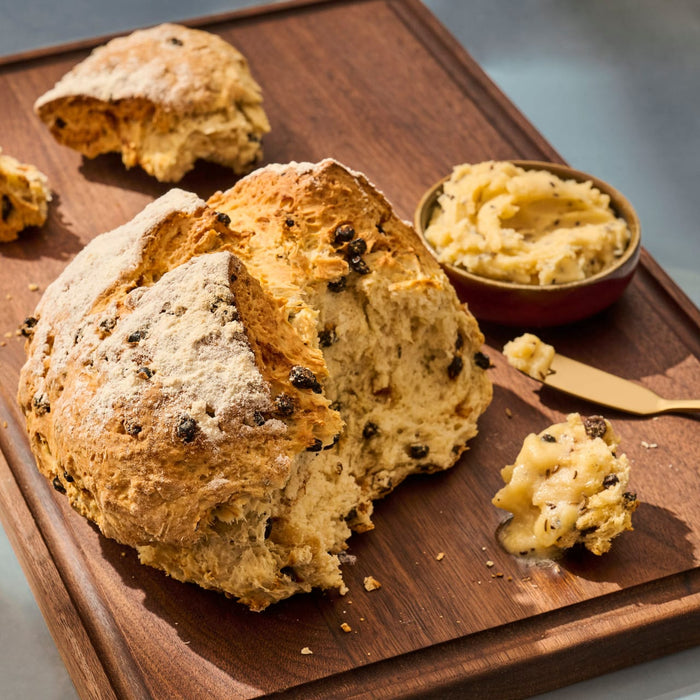 A loaf of Irish soda bread with currants is displayed on a wooden board, accompanied by a bowl of butter.