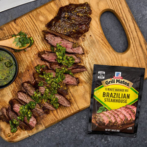 A sliced steak seasoned with McCormick Grill Mates Brazilian Steakhouse seasoning is displayed on a wooden cutting board with a side of green sauce.