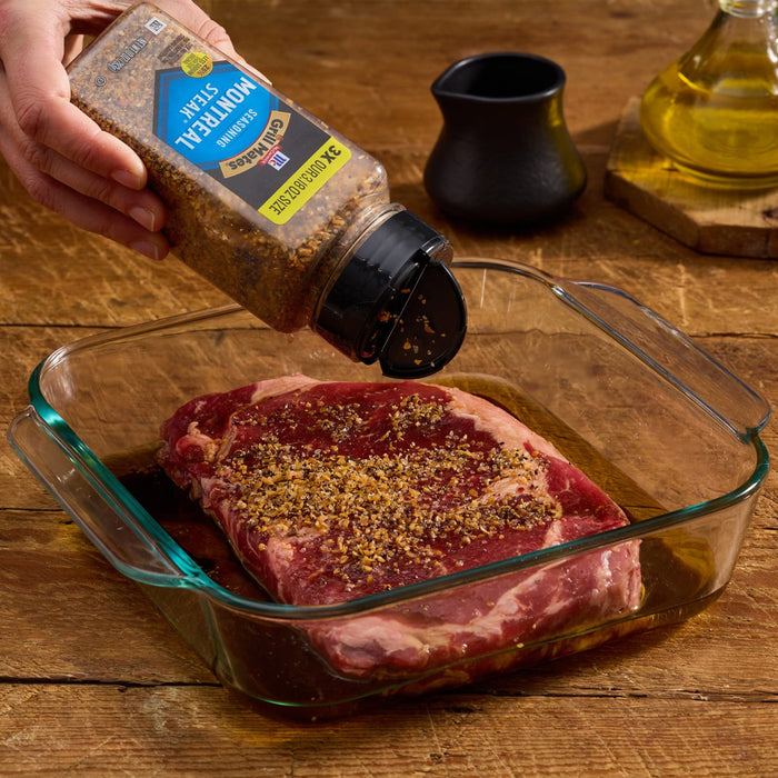 A hand sprinkles McCormick Grill Mates Montreal Steak Seasoning onto a raw steak in a glass dish.