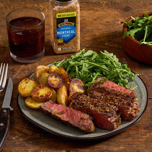 A plate of medium-rare steak crusted with McCormick Montreal Steak Seasoning, served with roasted potatoes and a side salad, next to a glass of dark liquid and the seasoning bottle.