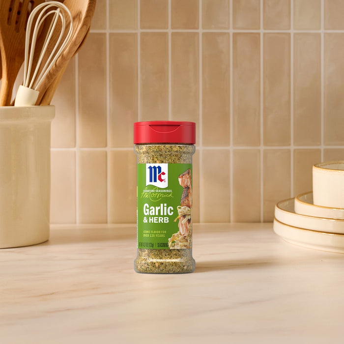 A bottle of Mccormick Garlic And Herb Pepper Seasoning Ground 4.37 Oz stands on a marble countertop in a bright kitchen setting with cooking utensils and stacked plates in the background.