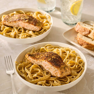 A delicious meal featuring two bowls of creamy pasta topped with pan-seared salmon fillets seasoned with Mccormick Garlic And Herb Pepper Seasoning Ground 4.37 Oz alongside sliced bread and lemon water.